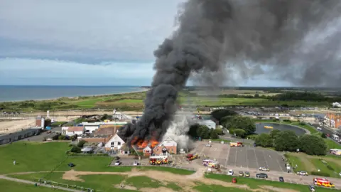 Eddie Mitchell A building with a plume of smoke and flames coming from the roof.