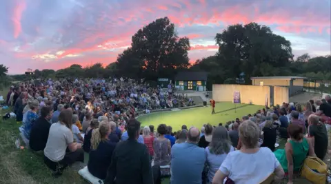 A pink cloudy sky at sunset is seen over an open air theatre where people sit on banked seating in a semi circle around a stage. The stage has a green floor and one performer on it wearing a red jacket.