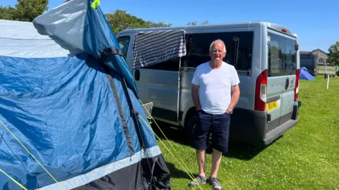A man with short white hair wearing a white t-shirt and dark blue shorts standing alongside a blue tent and van on a grassy field on a sunny day.