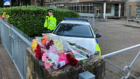 Tom Ingall/BBC Bunches of flowers are laid atop a wall. On the other side of the wall and adjoining gate is a police car, with a police officer standing beside the vehicle.