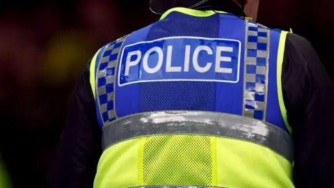 The back of a police officer, who is wearing a long-sleeved black top and a green and blue hi-vis jacket, that reads "police"