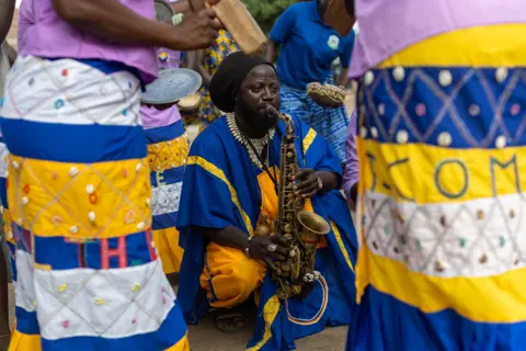 JEROME FAVRE / EPA A man crouches on the ground playing a saxophone. People dancing near him. They all wear blue and yellow clothing.