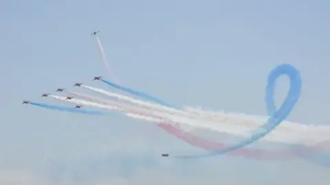 Nine aeroplanes in a line formation with two twirling in the sky with white blue and red smoke coming out behind them.