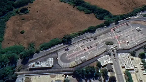 An aerial view of a road dividing an industrial campus and car park from a green area with a field and woodland.