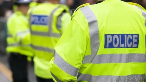 Three police officers standing outside, all with their backs to the camera. They are all wearing fluorescent yellow jackets with silver stripes across the backs and sleeves. On the back of the jackets there is a blue rectangle with the word, POLICE written inside.