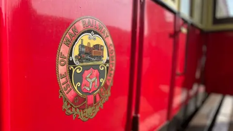 Side view of a red train carriage with the Isle of Man Railway Company logo featuring the Manx three legs of man symbol.