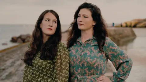 The Glasshouse The Unthanks which is made up of two women with dark, long hair. The woman on the left is looking at her sister over her shoulder. She is wearing a green patterned long-sleeved dress. The woman on the right is looking to the left and is wearing a blue and pink flowery dress. They are standing on a beach with the sea to the left.