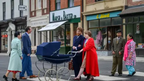 Men and women dressed in 1950s attire cross a road at the museum. One couple is pushing a pram with a South Asian couple walking behind them and another couple walking towards them. 