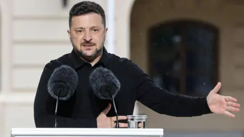 Volodymyr Zelensky stands at a white podium in front of a building. The podium has the Ukrainian flag on it and two microphones and the president is dressed in black and gesturing to the right side of the image with his hand