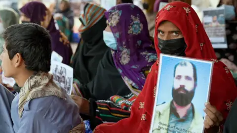 BBC/Nayyar Abbas A woman with a red head covering and a mask holds up a photo of a missing relative - she is sitting in a row of women, also holding photos.  