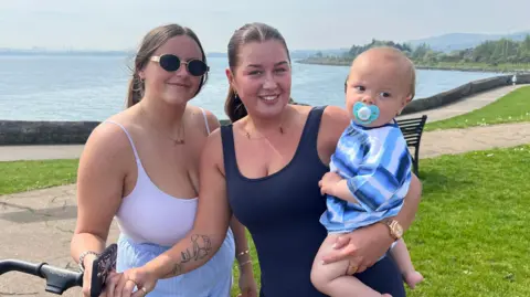 Two women stood side by side at the seafront. The woman on the left is wearing a white vest and has one hand on a child's buggy. The other woman is wearing a navy blue vest and is holding a child in her left arm. The baby has a dummy in its mouth and is wearing a blue striped shirt. 