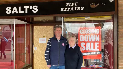 BBC A man wearing a blue gilet and striped shirt stands next to a woman with silver hair, glasses, and a dark jumper with a light collar. He has his arm around her and they both smile at the camera. They stand in front of a shop with red closing down signs in the window. The shop sign, which is black, says Salt's Pipe Repairs.