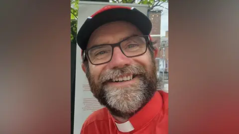 Supplied Chris Howson has slightly receding brown hair which is either short or tied back, and has a greying beard and black glasses. He is wearing a brown and green top, with a green shirt with white priest's dog collar. He has a neutral expression.