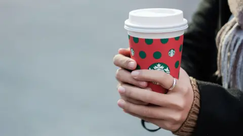 A woman holds a Christmas-themed Starbucks coffee cup. She's wearing a black coat, scarf and a silver ring on her left index finger.