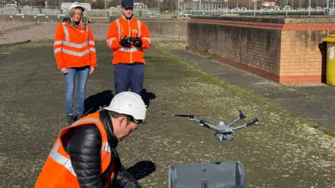 A drone is in the air above the lid of a grey box. The drone is grey with four "arms" for its rotors. A man kneels next to the box, with short dark hair, wearing an orange high-vis vest over a dark puffy jacket and a white hard hat. Behind him are a man and a woman. The woman has long blonde hair and wears an orange high-vis jacket, blue jeans and a white helmet. The man to her left has a blue baseball cap on his head, an orange high-vis jacket and blue trousers. He holds a black box in his hands.