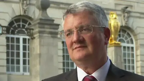 Leeds council leader James Lewis: a man with short grey hair and glasses. He is wearing a suit with a burgundy tie and is stood against the backdrop of Leeds Civic Hall.