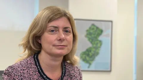 Lyndsay has shoulder-length blonde hair sits in an office chair wearing a pink and black textured jacket. Behind her are tall windows with blinds, a framed map on the wall, and a large potted plant.