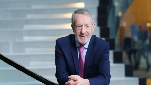 A grey short haired and bearded man wearing a navy suit and pink tie standing hands together on a bannister by some white stairs.