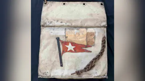 Henry Aldridge & Son The Titanic lifeboat seat cushion, including a piece of rope. It is a white cushion and has a red flag on it with a white star inside. 