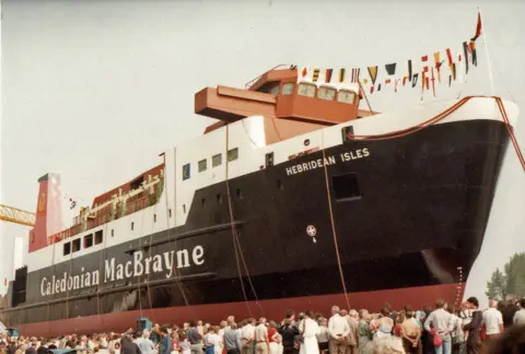 Brian Pulleyn Crowds gather in front of a large black and white ship with a red funnel as it is about to be launched