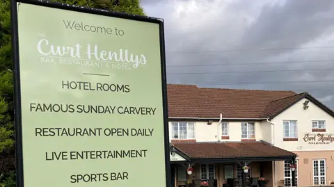 Cwrt Henllys Bar & Restaurant, a two-storey building painted cream and peach, with a red tile roof and a large tarmac car park. In the foreground, a sign advertises the restaurant's 'famous Sunday carvery'.