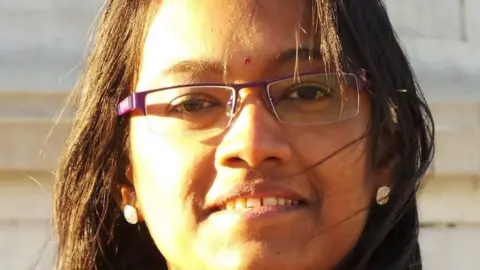 Valli wears glasses and has long dark hair and a small bindi on her forehead.