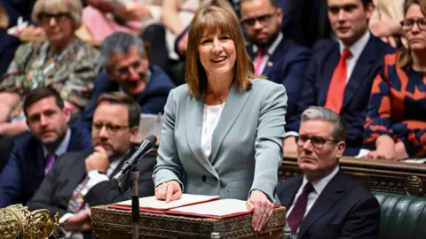 Rachel Reeves in a lightly coloured suit, pointing at her notes in a binder at the despatch box. Her party colleagues surround her, with Sir Keir Starmer sat closest to the right of the shot.