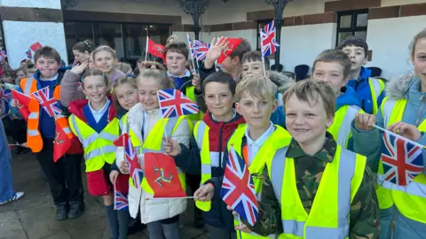 A group of primary school children wearing yellow high vis vests and holding Union Jack flags and Manx flags.