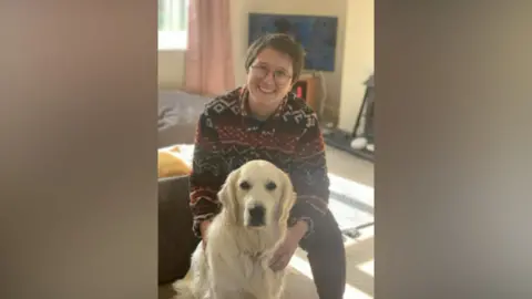 Family handout Ella Cook smiles at the camera as she squats on the floor with a white dog. They are both in a living room of a house. Ella has short brown hair, black rimmed glasses and is wearing a black and red fleece.