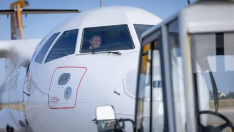 A first officer sits inside the cockpit of an Aurigny plane. He is smiling. A plane taxi is at the front of the photo as it pulls the plane, which has a yellow tail fin.