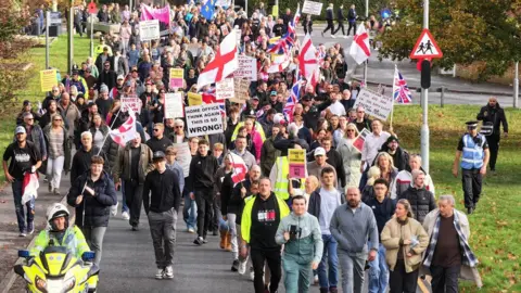 Eddie Mitchell Hundreds of people holding St George flags marching along a residential road.
