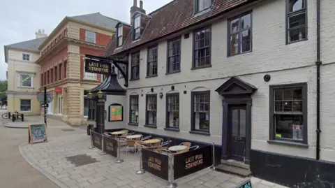 Google A pub with grey walls and dark grey window and door frames is shown with its name sign above the door to the left and an empty seating area in the centre of the frame.