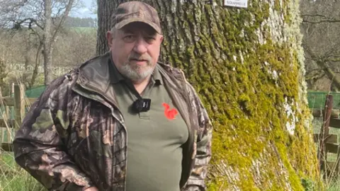 Ian Glendinning is a man in his 60s. He is standing in front of a tree wearing a camouflage jacket and a green top with a red squirrel logo on it.