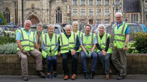 Eight people sit on a low stone wall as they pose for a picture. They're all wearing florescent yellow jackets. 