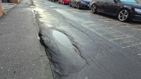 A very large pothole on a residential street. The hole has a puddle of water in it, and is so severe that some of the tarmac has been pushed up onto the pavement. 