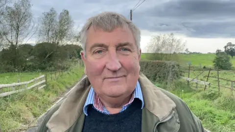 County councillor James Elson stands on road in front of destroyed Llanerch bridge, wearing a green jacket, blue and red checked shirt and navy blue sweater