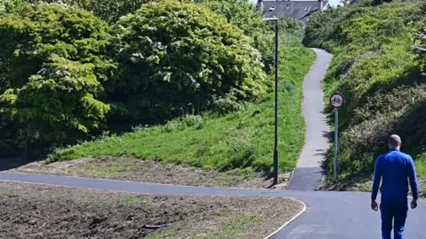 Cycle path through green hills with a red circle sign showing a cycle and a street light on the path. There is a man with his back to the camera walking the path. He is wearing a blue top and jeans.