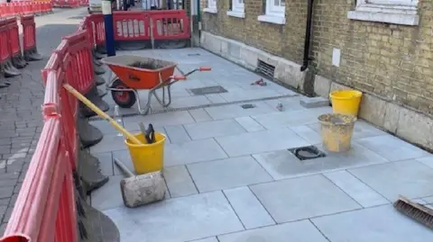 Cambridgeshire & Peterborough Combined Authority A wheelbarrow and builders items left on the ground for pavement work to be carried out. You can see three buckets, a red wheelbarrow, clean paving and red barriers. There is a brick wall to the right. 