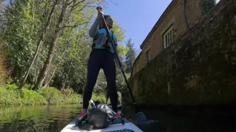 Sarah stands on her paddleboard, navigating her way down the river. The picture is taken from a miniature camera at the tip of her board, looking upwards at her.