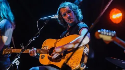 Kristen Nickel A man is playing a guitar on a stage. He is sitting on a chair and has a tall microphone in front of him. He has shoulder length dark hair. 