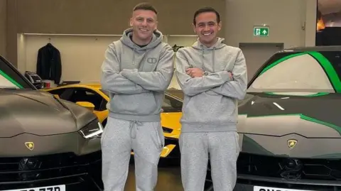Two people in matching grey tracksuits stand with arms folded between two Lamborghini SUVs in a showroom, with a yellow sports car visible behind them and UK number plates on the vehicles.