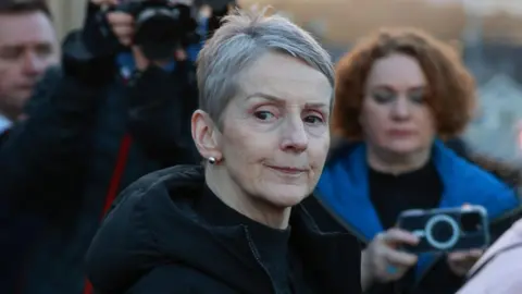 A close-up image of Lady Eleanor Donaldson as she walks out of court. She is an older woman with short, grey hair and has a neutral expression on her face. There are journalists in the background taking videos and pictures of her. Lady Donaldson is wearing a black, puffer raincoat and a pair of silver stud earrings.