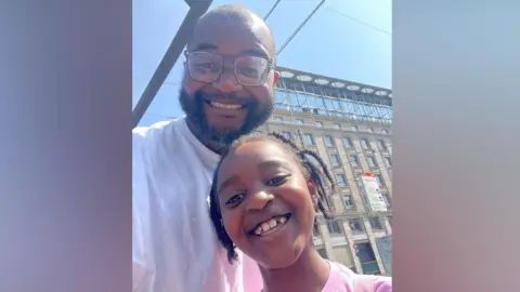 Anthony Ojo and his daughter. Anthony is bald and is wearing glasses and a white t-shirt. His daughter is wearing a pink t-shirt and has braids. Both are smiling to camera.