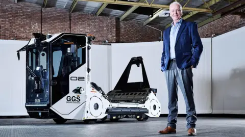 David is wearing a mid-blue blazer over a light blue shirt and light grey trousers. He is standing in a warehouse and in front of an autonomous baggage handling vehicle, made by his firm.