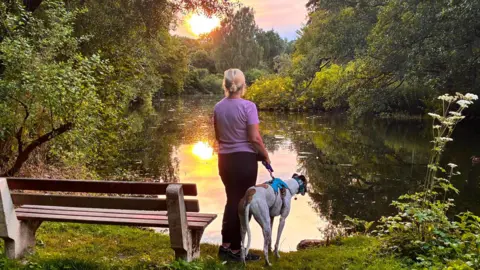 Battersea Dogs and Cats Home A woman in black trousers and a pink top holding a greyhound on a lead, while looking out across a pond at sunset