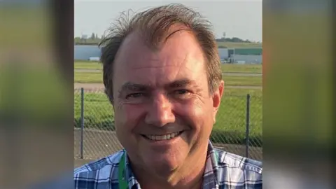 A man in a black and white shirt smiling at the camera. They are standing in front of a green field with buildings on it.