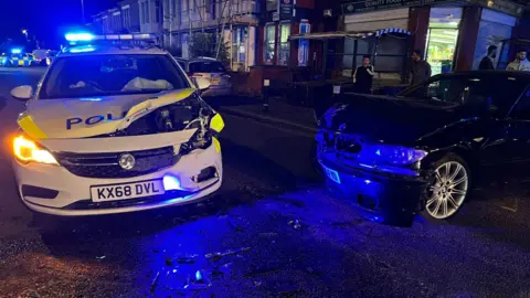 A damaged police car and BMW are illuminated in the night by the blue lights of the police car, which has its airbags engaged