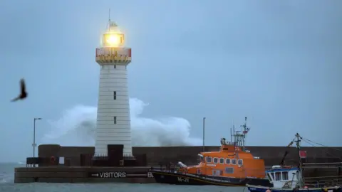 A wide shot of boats in front of a pier with a lighthouse on it. A wave can be seen striking the lighthouse from behind. The sky is grey. A bird can be seen.