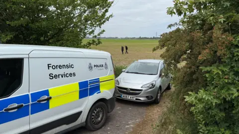 Steve Hubbard/BBC A police van and a car at the entrance to a field. The van says 'forensic services' on it. In the background, there are two people in dark clothing walking across the field.