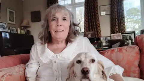 Supplied Geraldine at home with her white dog Alice. They are sitting together on a couch. Geraldine is wearing a white blouse and has shoulder length grey hair that is curly at the ends, she is smiling at the camera. Behind her are lots of framed family photos.